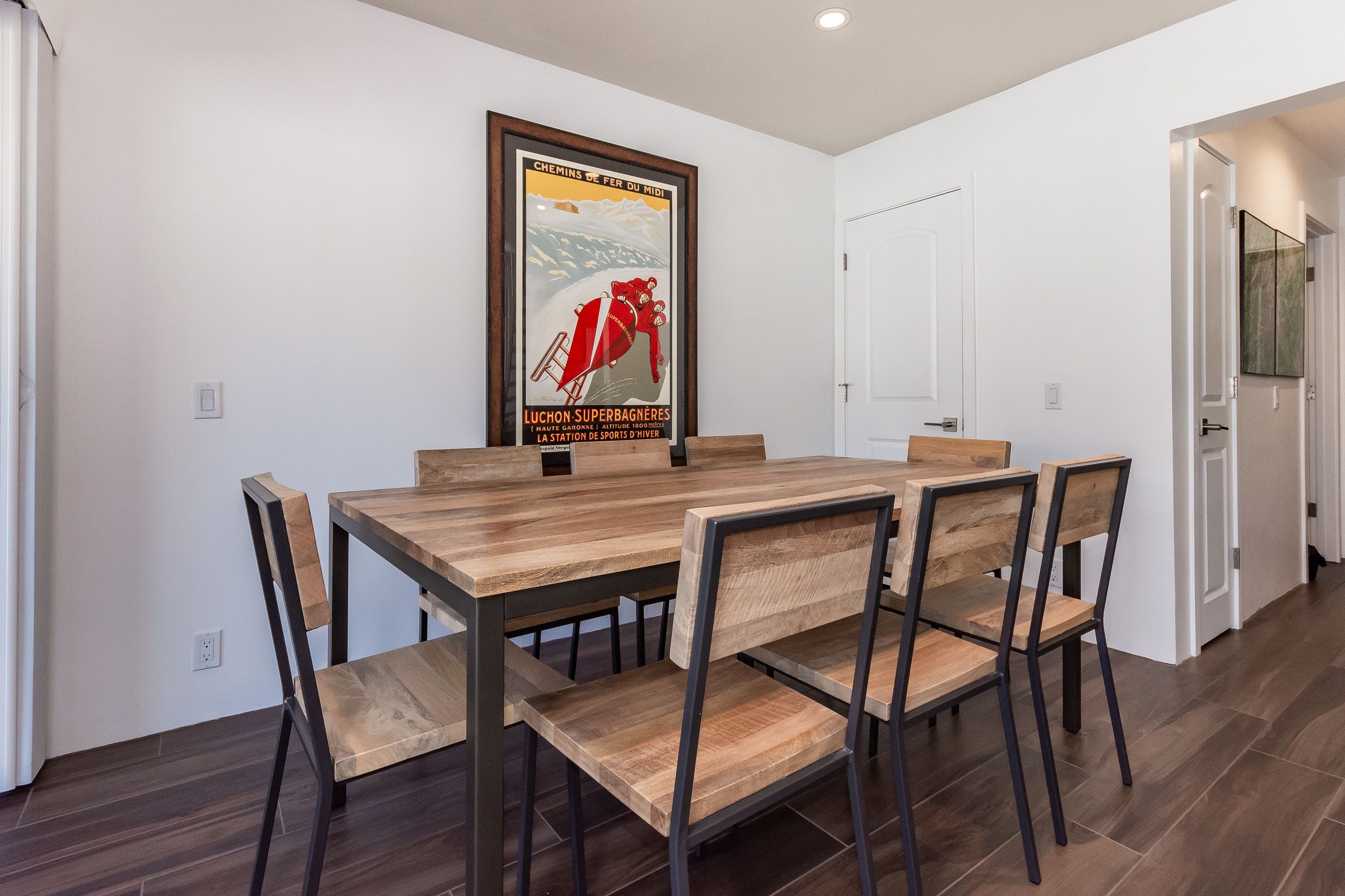 Uma sala de jantar com uma mesa e cadeiras de madeira, um cartaz emoldurado na parede e uma porta branca ao fundo.