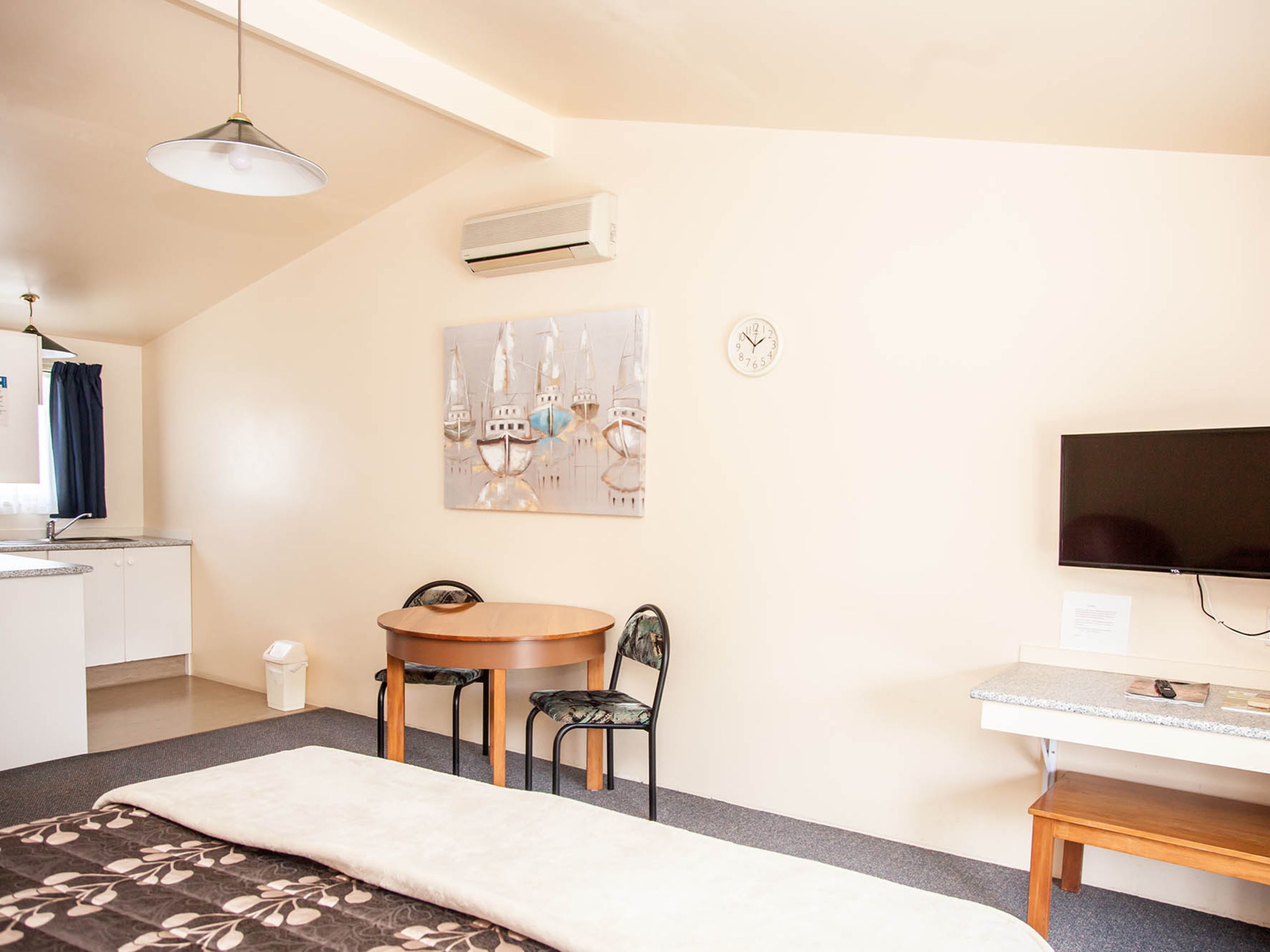A compact room with a bed, a small table with two chairs, a TV, an air conditioner, and a painting on the wall.