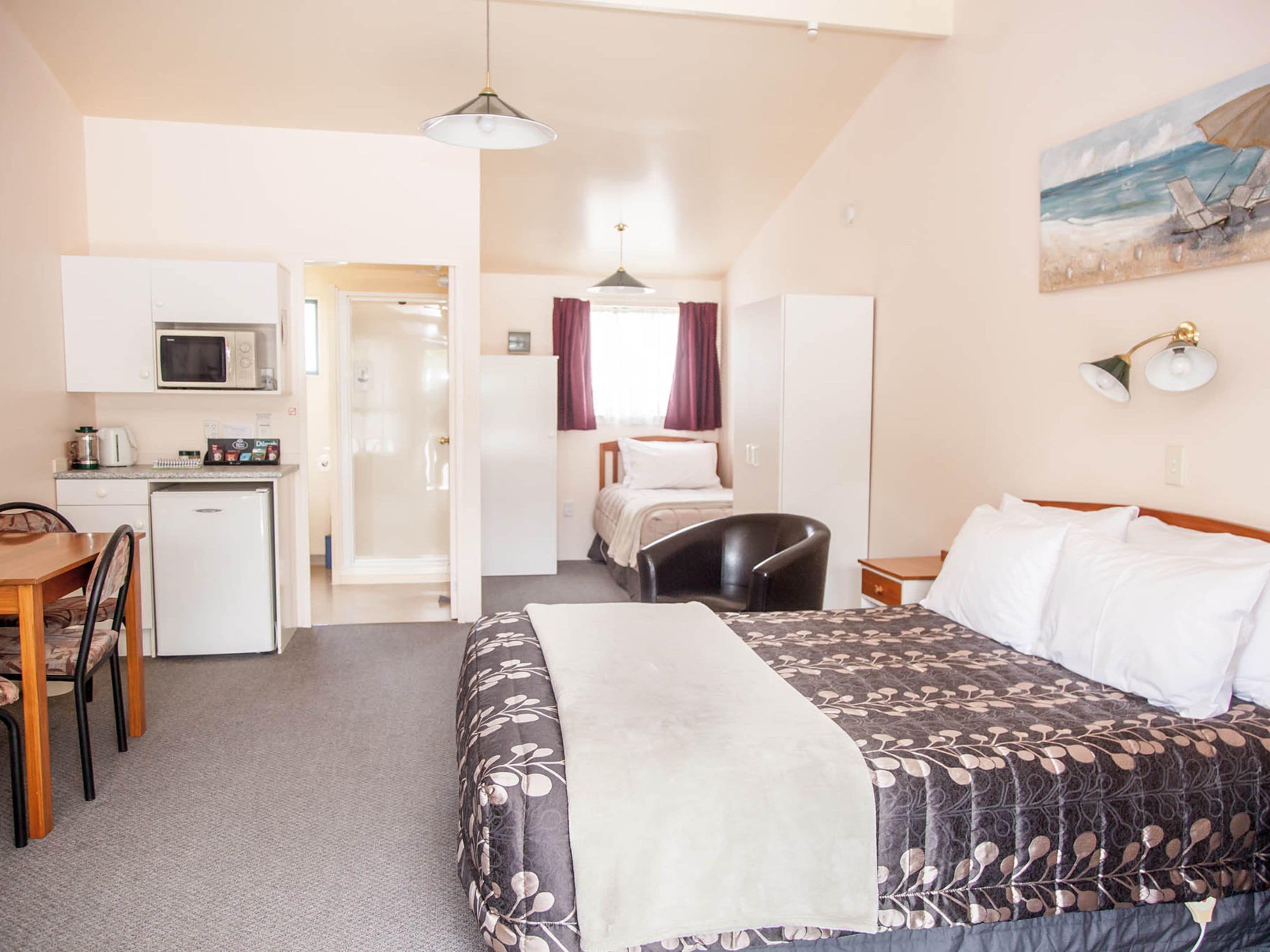 A compact hotel room with a bed, a desk with a microwave, a refrigerator, a chair, and a painting on the wall.