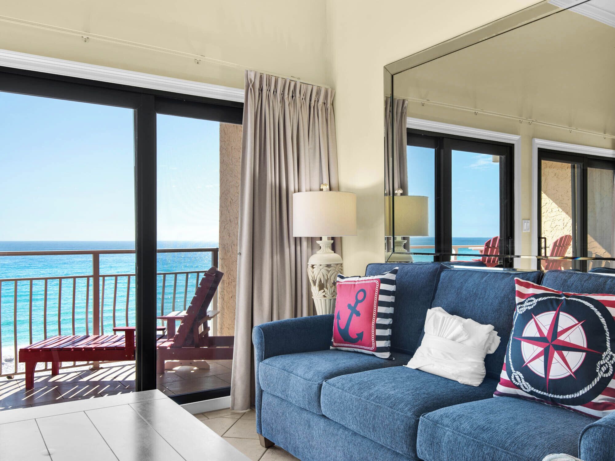 A balcony with a view of the ocean, a blue sofa with nautical-themed pillows, and a red chair.