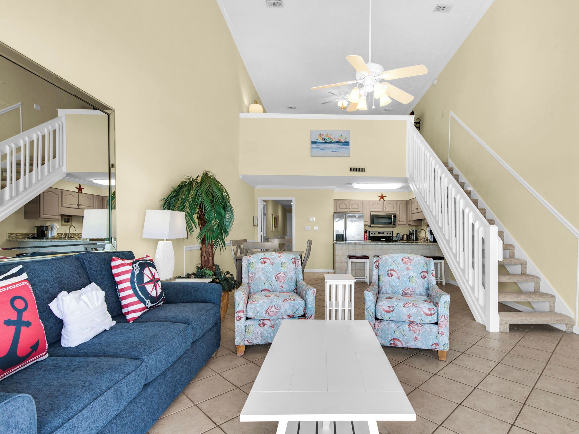 A spacious living room with a blue sofa, two patterned armchairs, a white coffee table, and a ceiling fan. A staircase leads to an upper level with a kitchen and dining area.