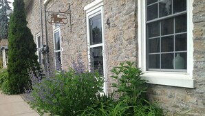 Un bâtiment en pierre avec un cadre de fenêtre blanc, une enseigne suspendue représentant un lion, et un jardin bien entretenu avec des plantes vertes et des fleurs violettes.