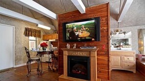 Ein Wohnzimmer mit Kamin, einem darüber angebrachten Fernseher und einer Essecke mit Holztischen und -stühlen.