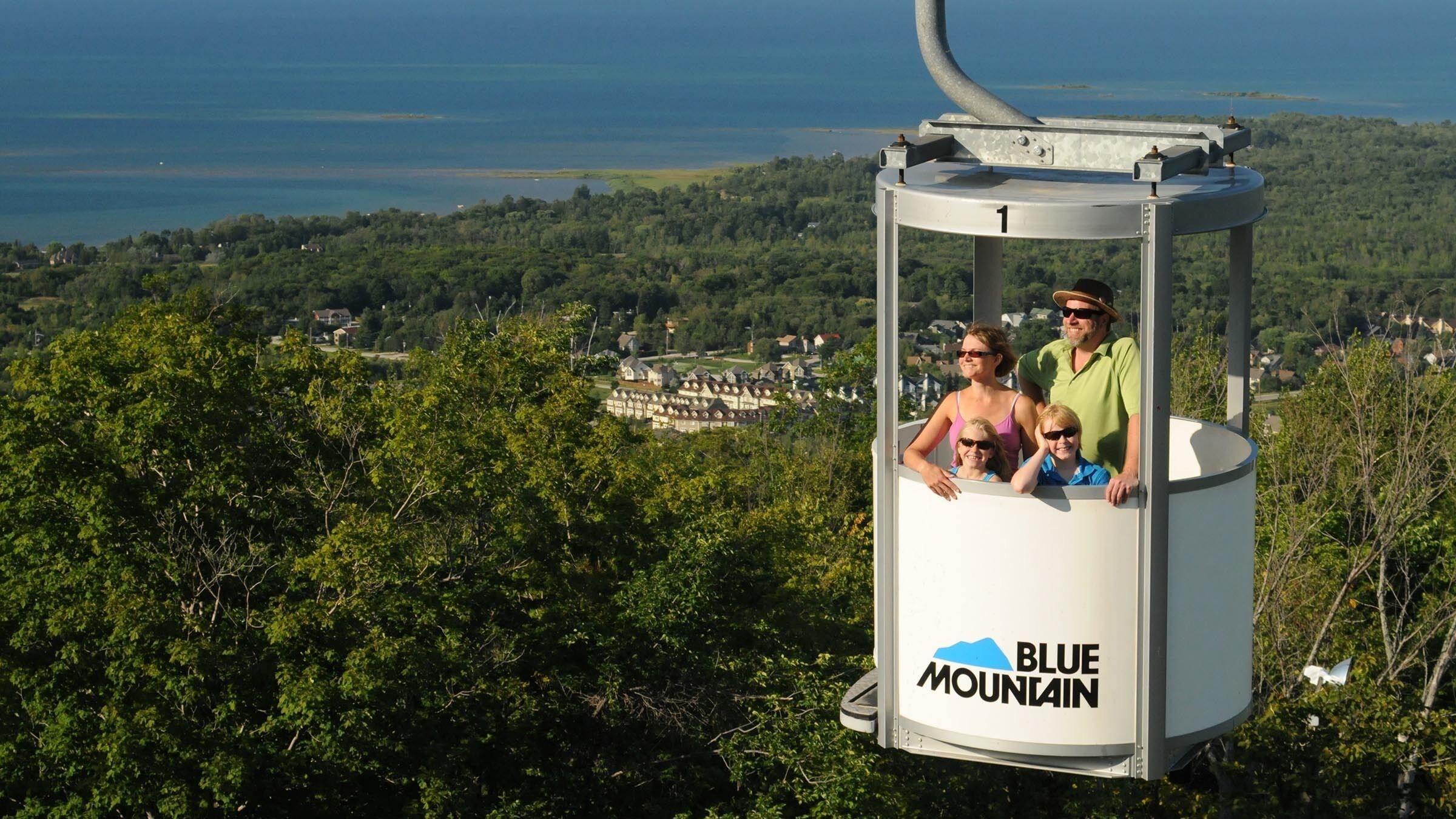 Une télécabine arborant le logo « Blue Mountain », avec des passagers à l’intérieur, et une vue sur une ville et une zone boisée.