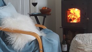 A cozy room with a chair covered in a blue blanket, a white fluffy pillow, a small table with a wine glass and a bowl, and a wood-burning stove. - Mapua Studio  High Quality Accommodation  (Mapua (Ruby Bay))