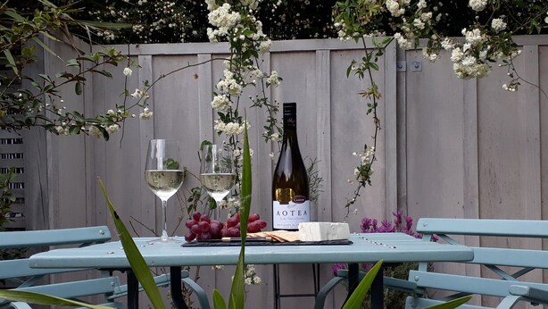 A table set with a bottle of wine, two glasses, and a cheese plate, surrounded by greenery and flowers. - Mapua Studio  High Quality Accommodation  (Mapua (Ruby Bay))