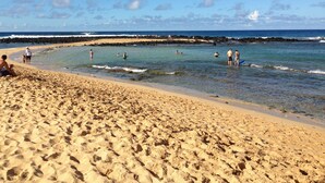 Una playa arenosa con gente nadando y de pie en el agua, y una costa rocosa al fondo.