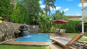 Una piscina con una pared de piedra, una cascada y un patio con sillones de descanso y un paraguas rojo.