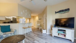 A living room with a sofa, chairs, a coffee table, a TV, and a kitchen area. - 48 Ocean Breeze: Pet Friendly, Pool Views, Free Bikes, Tennis, Walk to Beach (Hilton Head Island)