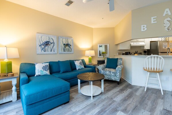 Ein modernes Wohnzimmer mit einer Couch, einem Sessel und einem runden Couchtisch. Im Hintergrund ist eine Küchenzeile mit einem Kühlschrank und Schränken zu sehen.