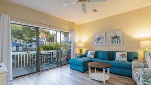 A living room with a blue sofa, a round coffee table, and a balcony with a glass door. - 48 Ocean Breeze: Pet Friendly, Pool Views, Free Bikes, Tennis, Walk to Beach (Hilton Head Island)