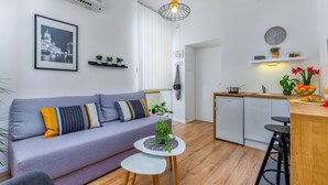 En moderne lejlighed med en grå sofa, et lille rundt bord med en plante, et køkken med træbordplade og et spiseområde med en barstol.