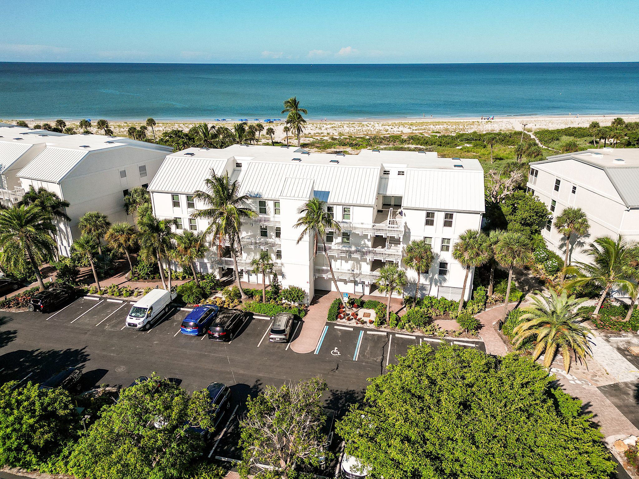 A multi-story hotel with a parking lot, surrounded by palm trees and a view of the beach.