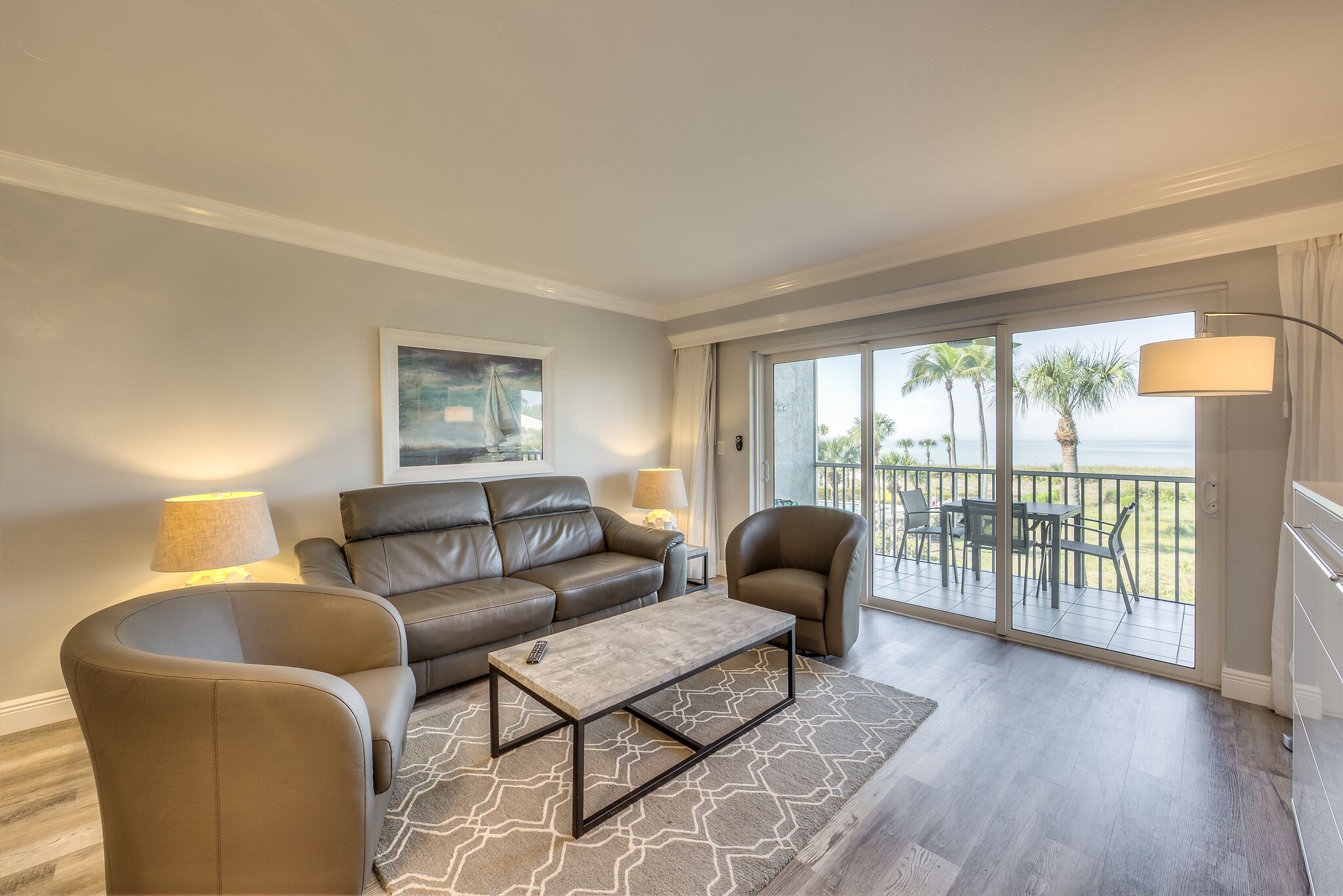 A modern living room with a sofa, armchair, and coffee table, a painting on the wall, and a balcony with a view of palm trees and the sea.