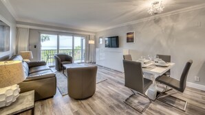 A modern living room with a sofa, ottoman, and dining table set for six. A TV is mounted on the wall, and there is a balcony with a view of the ocean. - Stunning Modern beach front unit - completely renovated!  Includes golf cart! (Captiva)