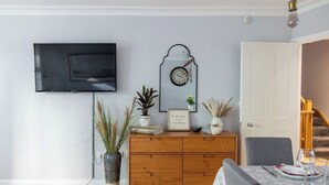 A living room with a flat-screen TV mounted on the wall, a wooden cabinet with decorative items, a dining table set for two, and a staircase leading to another level. - Spacious quiet 4BR/6beds house with private garage (London)