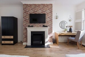 A living room with a brick fireplace, a flat-screen TV mounted above it, a wooden desk with a chair, and a black wardrobe.