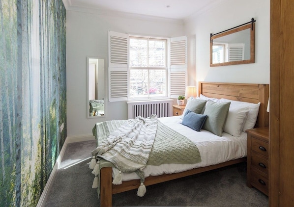 A bedroom with a bed, a nightstand, a mirror, and a window with shutters.