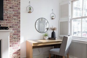 A room with a brick wall, a wooden desk, a chair, a mirror, and a window with blinds.