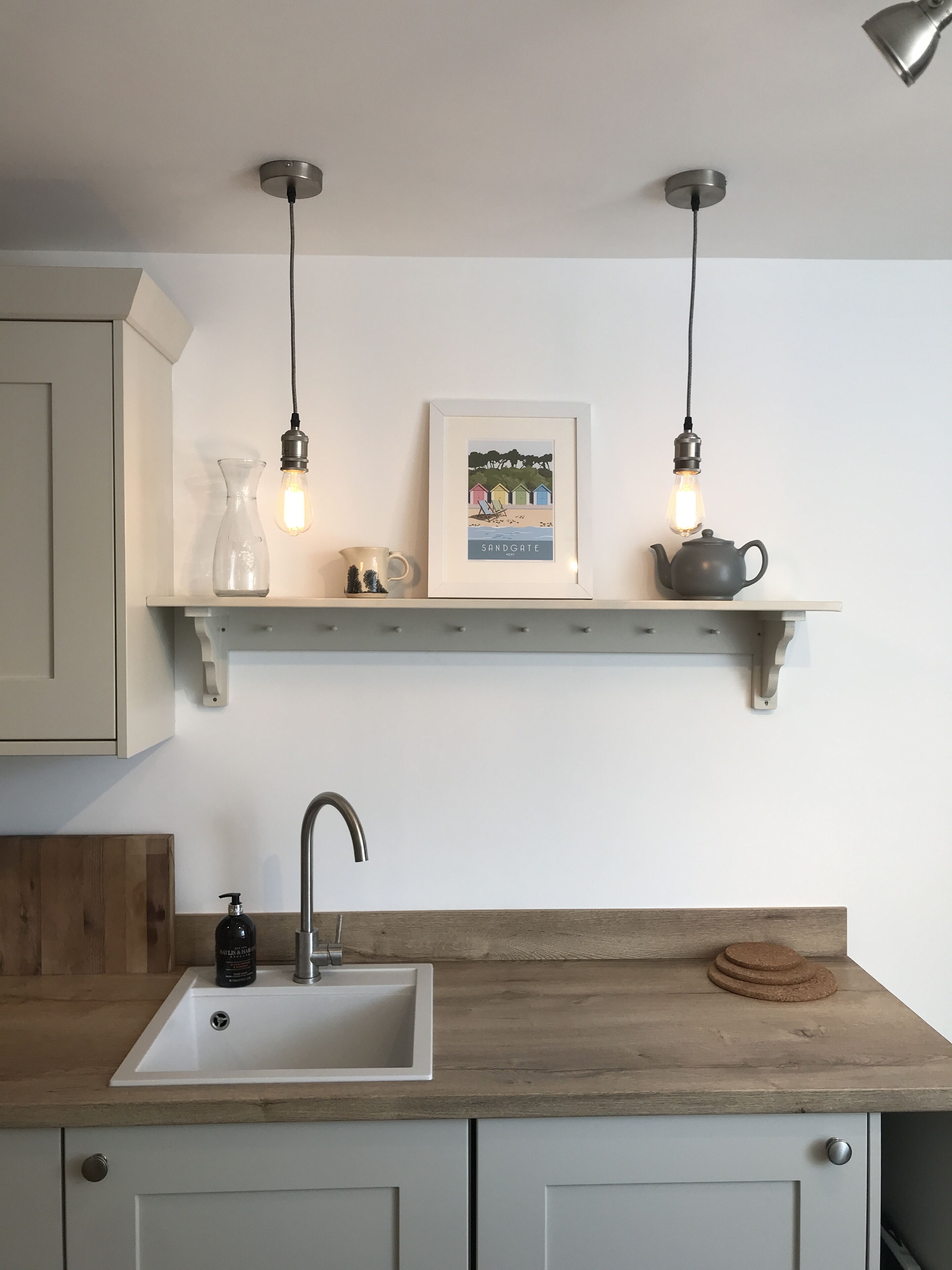 Eine Küche mit einem weißen Spülbecken, einer Holzausziehtafel und einem Regal darüber, auf dem sich ein gerahmtes Bild, ein Teekessel und eine Glasvase befinden.