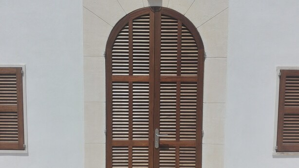 Une grande porte en bois, composée de lattes verticales, flanquée de deux fenêtres dotées de volets en bois.