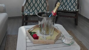 Une petite pièce bien éclairée, avec une table blanche, deux chaises à motifs, un canapé et une fenêtre avec des stores.