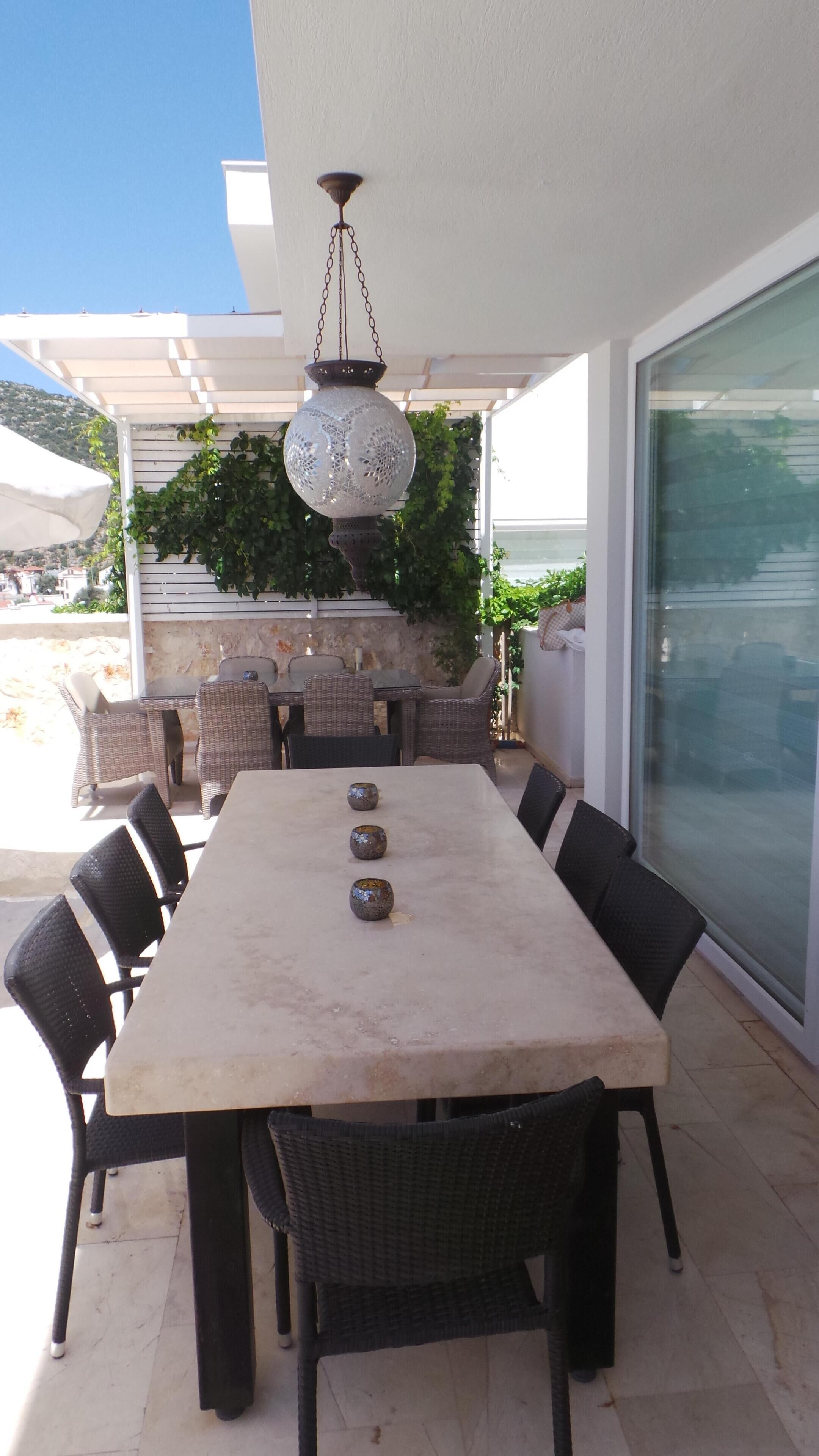 An outdoor dining area with a large table and chairs, a hanging light fixture, and a view of the surrounding landscape.