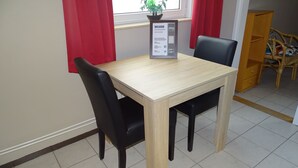 Un área de comedor con una mesa de madera y sillas negras, un letrero de bienvenida, una ventana con cortinas rojas y una silla de mimbre al fondo.