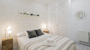 A neatly arranged bedroom with a bed, bedside lamps, a shelf with plants, a radiator, and a mirror on the wall.