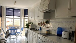 Una cocina moderna con armarios blancos, horno empotrado y un fregadero. Hay una zona de comedor con sillas azules y una mesa.