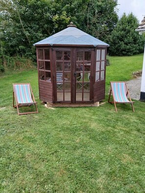Ein hölzerner Pavillon mit Glastür und Fenstern, umgeben von einer grünen Rasenfläche und zwei gestreiften Liegestühlen.