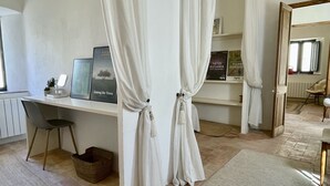 A room with a desk, chair, and shelves with books. There is a window with curtains, a radiator, and a wicker basket. - CASA ROSA DEL PORT (L'Escala)