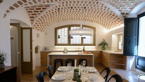 A dining area with a long table set for six, a kitchen in the background, and a brick ceiling. - CASA ROSA DEL PORT (L'Escala)
