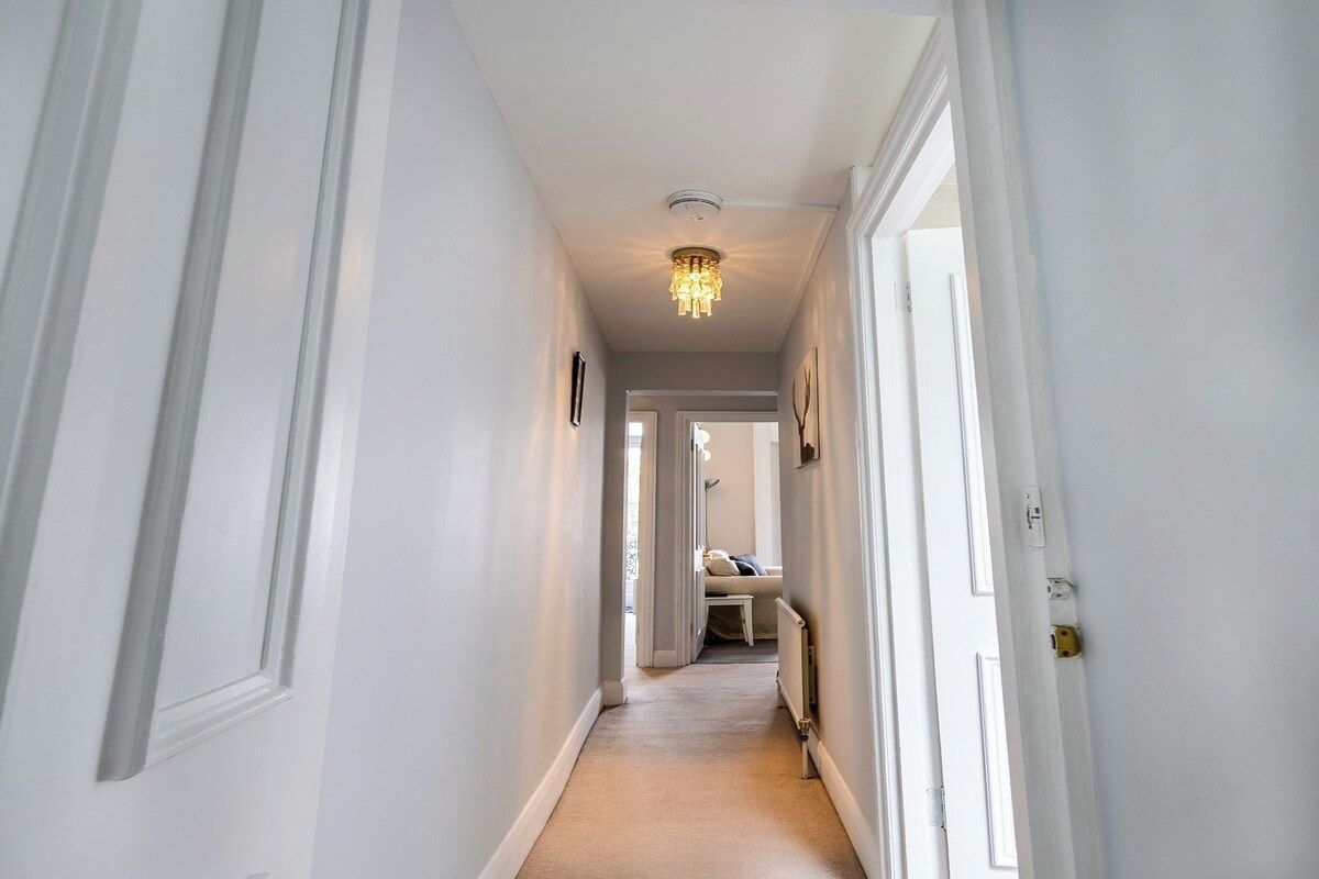 A hallway with white walls, a carpeted floor, and a chandelier. There are doors on both sides, and a bench in the distance.