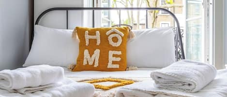 A neatly made bed with folded white towels, a yellow pillow with 'HOME' spelled out, and a window with a view of buildings and trees.