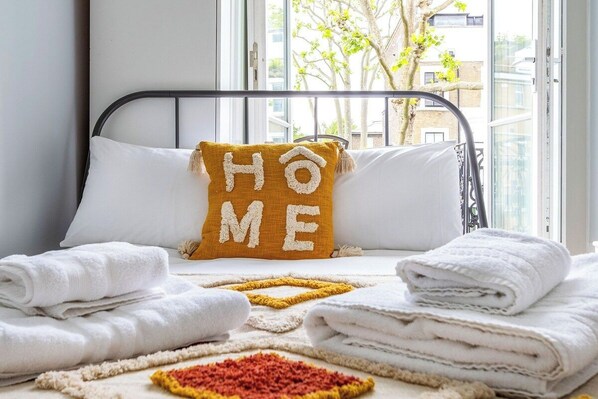 A neatly made bed with folded white towels, a yellow pillow with 'HOME' spelled out, and a window with a view of buildings and trees.