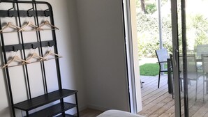 Un dormitorio con una cama, una estantería metálica negra con perchas de madera, una lámpara blanca y un ventanal corredizo que da a un patio exterior con una mesa y sillas.