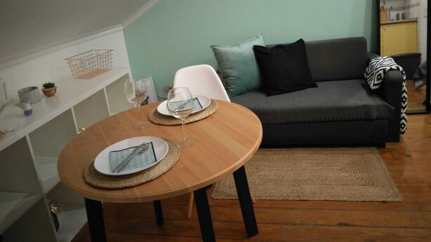 Ein gemütliches Wohnzimmer mit einem runden Holzes-Esstisch für zwei Personen, einem grauen Sofa mit Kissen und einer kleinen Vitrine mit Dekoration.