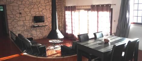 A dining area with a stone wall, a fireplace, a TV mounted on the wall, and a window with curtains.