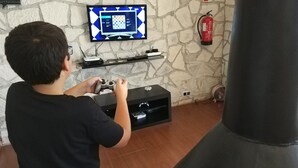 A person playing a video game in a room with a stone wall, a mounted television, a fire extinguisher, and a small shelf with electronic devices. - Louro's Villa in Vila Nova (Coimbra)