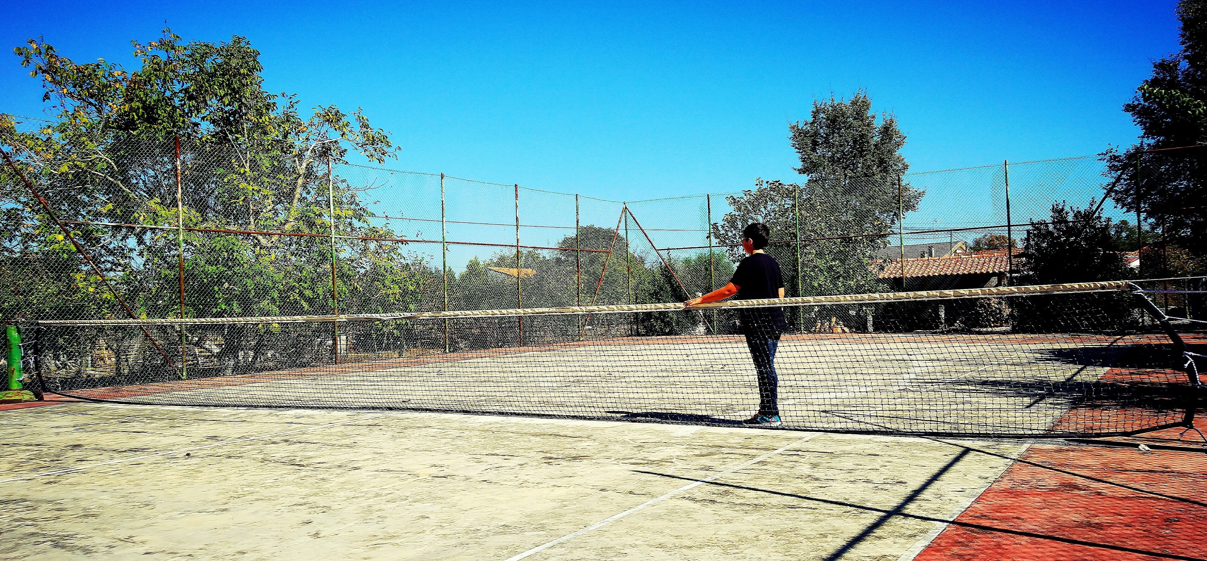 Een tennisbaan met net, omgeven door een hek en bomen.