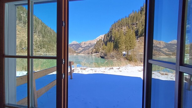 A room with a large window offering a view of a snowy landscape, a wooden deck, and a mountain backdrop.