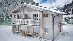 A two-story wooden cabin with a balcony, large windows, and a view of a lake and mountains.
