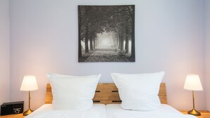 A neatly made bed with white linens, two bedside lamps, and a wall-mounted picture of a tree-lined pathway. - Frida - Family vacation home, WLAN free, sauna, fireplace, 8 persons (Fehmarn)