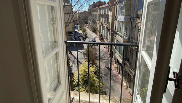 Un balcó amb vista a un carrer estret flanquejat per edificis, un cel serè i un arbre.