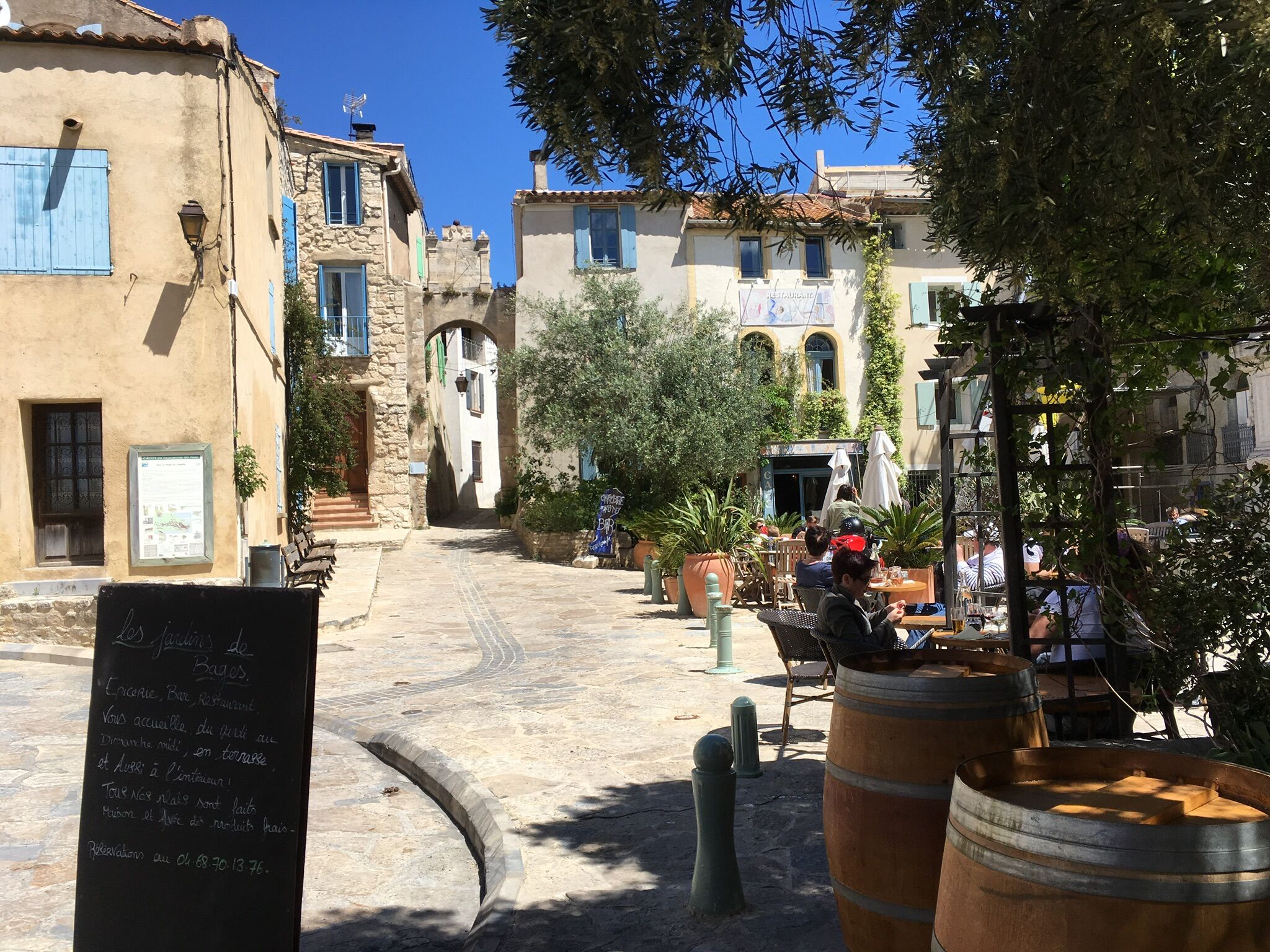 Une rue étroite bordée de bâtiments des deux côtés, avec des tables et des chaises à l’extérieur d’un café, et une enseigne avec du texte.