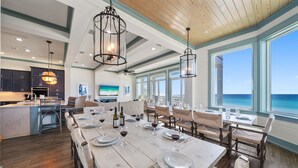 Un vaste espace de salle à manger avec une longue table dressée pour le repas, de grandes fenêtres offrant une vue sur l’océan et, au fond, une cuisine moderne.