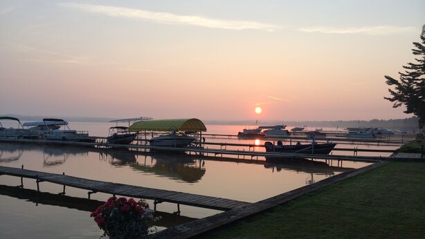 A marina with boats docked, a grassy area with a bench, and a sunset in the background. - Home on the lake. Walk out your door for Swimming, boating or morning coffee. (Mears)