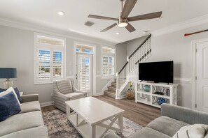 A living room with a ceiling fan, a TV on a white stand, a staircase, and a view of the outside through large windows.