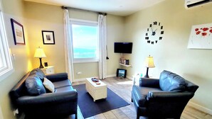 A living room with two black leather sofas, a white coffee table, a flat-screen TV, and a large window with curtains.